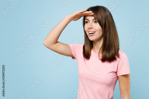 Young smiling happy fun caucasian woman 20s wearing pink t-shirt hold hand at forehead look far away distance isolated on pastel plain light blue background studio portrait. People lifestyle concept.