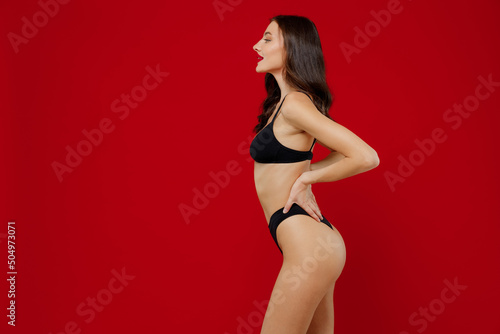 Side profile view young sexy brunette woman 20s with perfect fit body in black underwear hold hand on waist stand akimbo isolated on plain red background studio portrait. People female beauty concept
