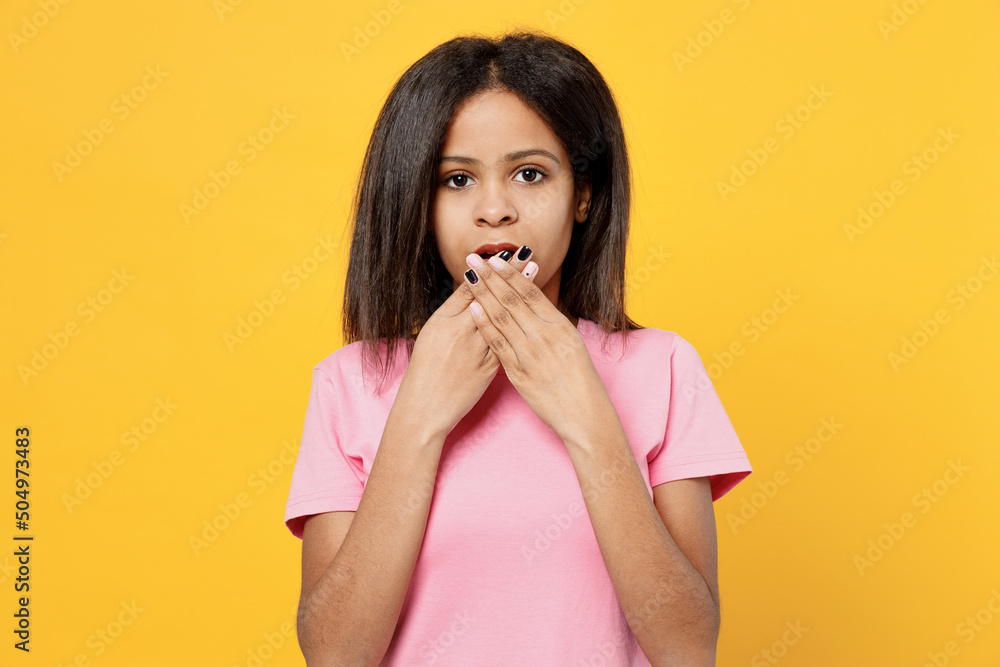 Little shocked surprised kid girl of African American ethnicity 12-13 years old in pink t-shirt cover mouth with hand isolated on plain yellow background studio portrait. Childhood lifestyle concept. Little shocked surprised kid girl of African American ethnicity 12-13 years old in pink t-shirt cover mouth with hand isolated on plain yellow background studio portrait. Childhood lifestyle concept.