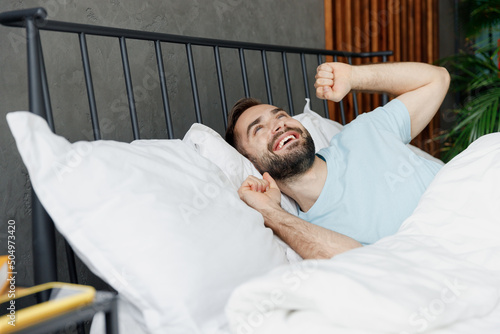 Young happy calm smiling fun cool man 20s in casual blue t-shirt lying in bed sretch hands rest relax spend time in bedroom lounge home in own room house wake up dream Good mood night bedtime concept photo