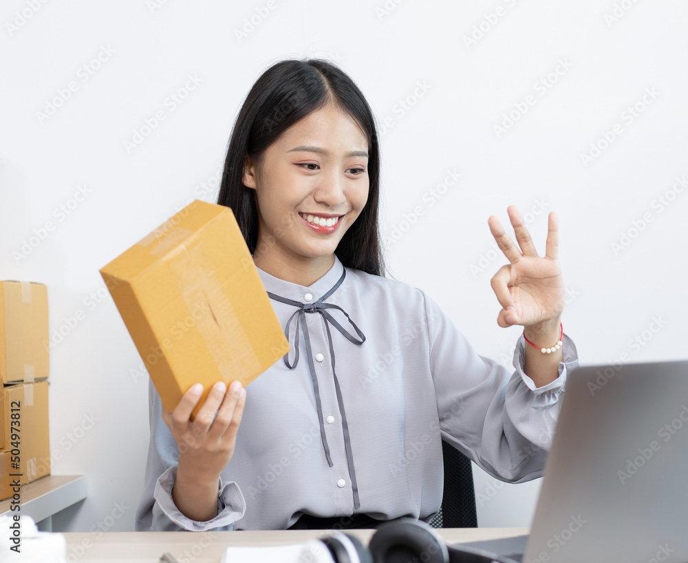 Young Asian woman chatting with a customer on a laptop and displays the product before delivery to confirm the order, Selling products online or doing freelance work at home concept. Young Asian woman chatting with a customer on a laptop and displays the product before delivery to confirm the order, Selling products online or doing freelance work at home concept.