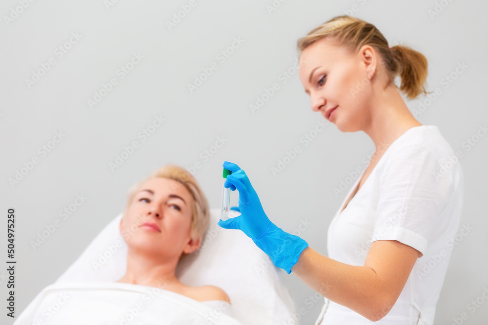 A female cosmetologist in rubber gloves shows a test tube to a client. A woman lying on a couch in defocus. Copy space. The concept of plasmolifting