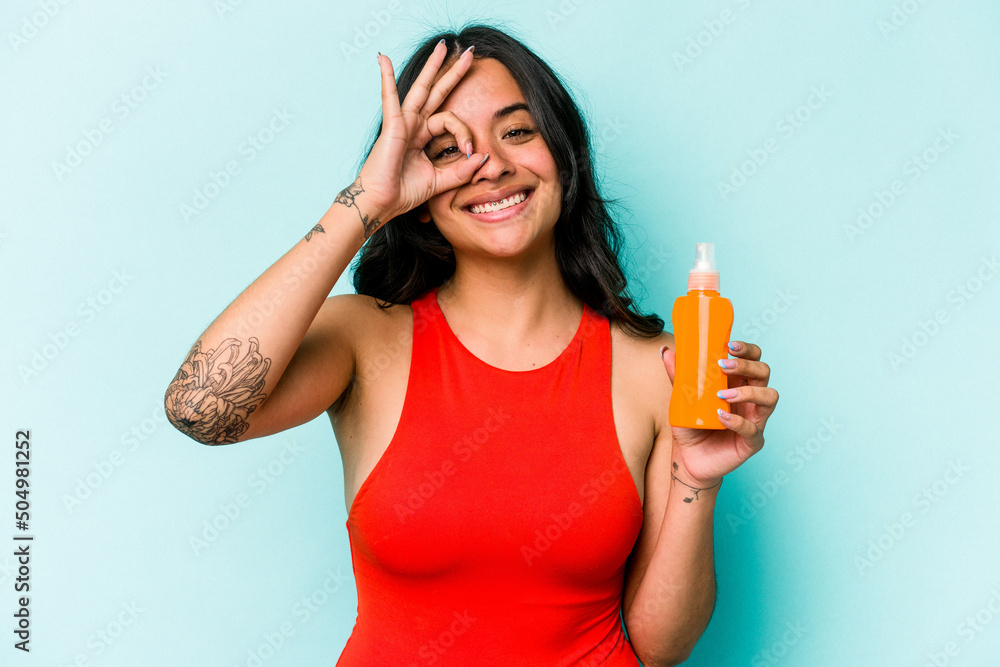 Young hispanic woman holding sun cream isolated on blue background excited keeping ok gesture on eye. Young hispanic woman holding sun cream isolated on blue background excited keeping ok gesture on eye.