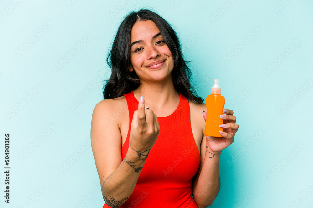 Young hispanic woman holding sun cream isolated on blue background pointing with finger at you as if inviting come closer. Young hispanic woman holding sun cream isolated on blue background pointing with finger at you as if inviting come closer.