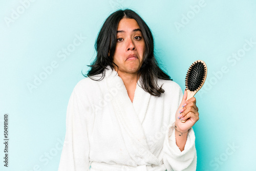 Young hispanic woman holding hairbrush isolated on blue background shrugs shoulders and open eyes confused.