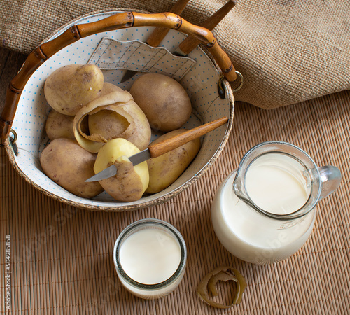 Vegan potato milk in a jug and glass and white raw potatoes in a wicker basket with a bamboo handle on a bamboo stand. Natural background. Making potato milk 