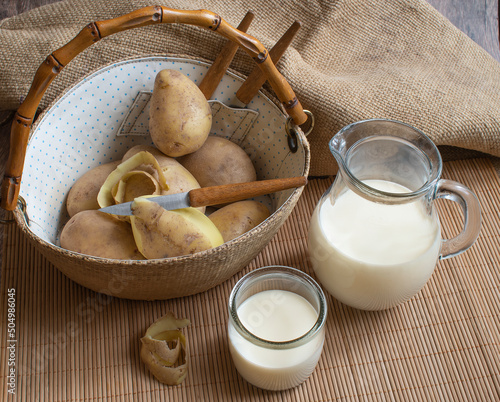 Vegan potato milk in a jug and glass and white raw potatoes in a wicker basket with a bamboo handle on a bamboo stand. Natural background. Making potato milk 