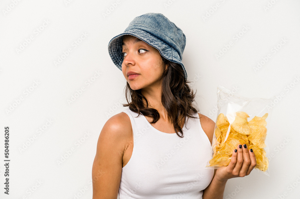 Young hispanic woman holding a bag of chips isolated on white background looks aside smiling, cheerful and pleasant. Young hispanic woman holding a bag of chips isolated on white background looks aside smiling, cheerful and pleasant.