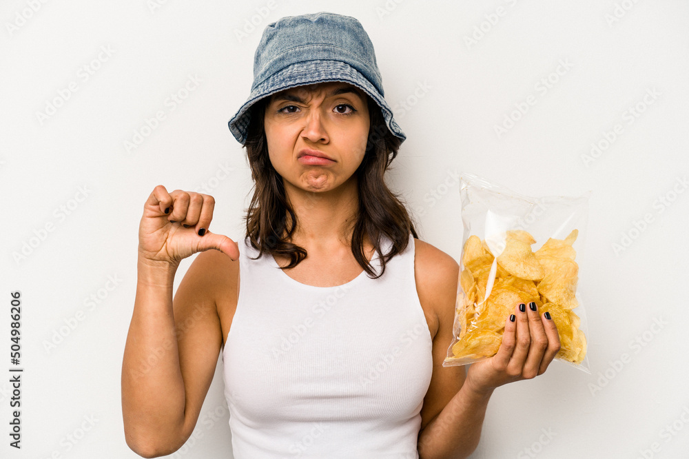 Young hispanic woman holding a bag of chips isolated on white background showing a dislike gesture, thumbs down. Disagreement concept. Young hispanic woman holding a bag of chips isolated on white background showing a dislike gesture, thumbs down. Disagreement concept.