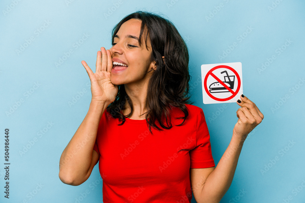 Young hispanic woman holding no eating sign isolated on blue background shouting and holding palm near opened mouth. Young hispanic woman holding no eating sign isolated on blue background shouting and holding palm near opened mouth.