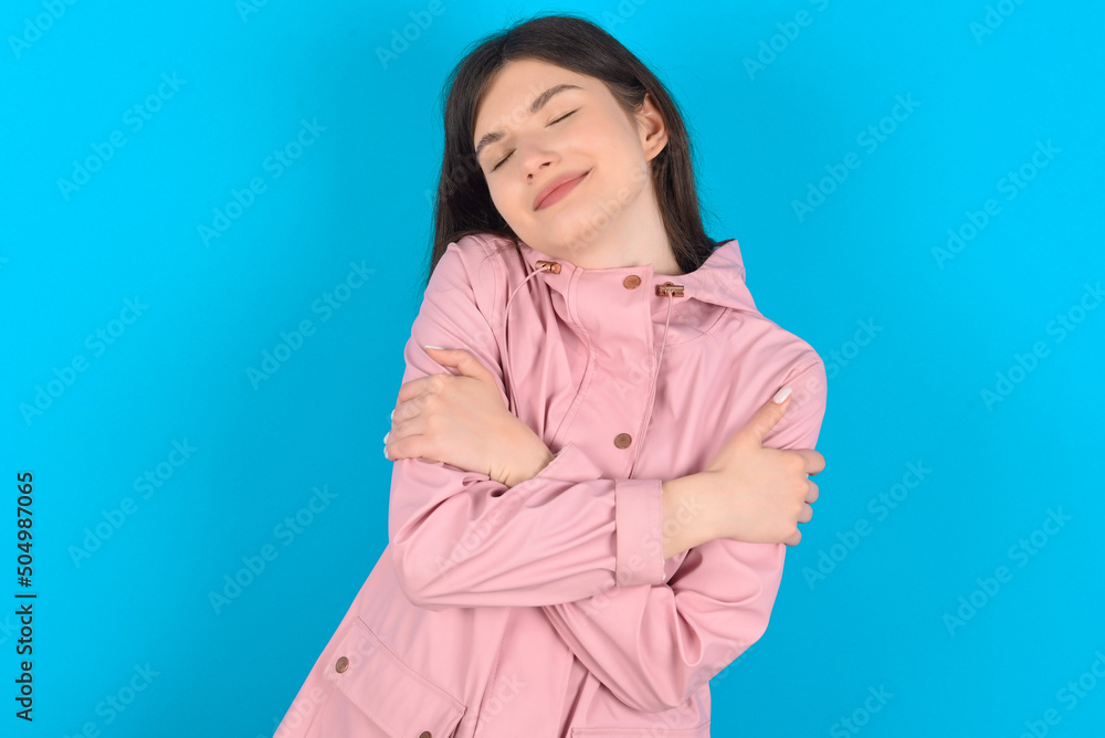 young beautiful Caucasian woman wearing pink raincoat over blue wall Hugging oneself happy and positive, smiling confident. Self love and self care young beautiful Caucasian woman wearing pink raincoat over blue wall Hugging oneself happy and positive, smiling confident. Self love and self care