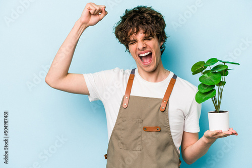 Young gardener caucasian man holding a plant isolated on blue background raising fist after a victory, winner concept.