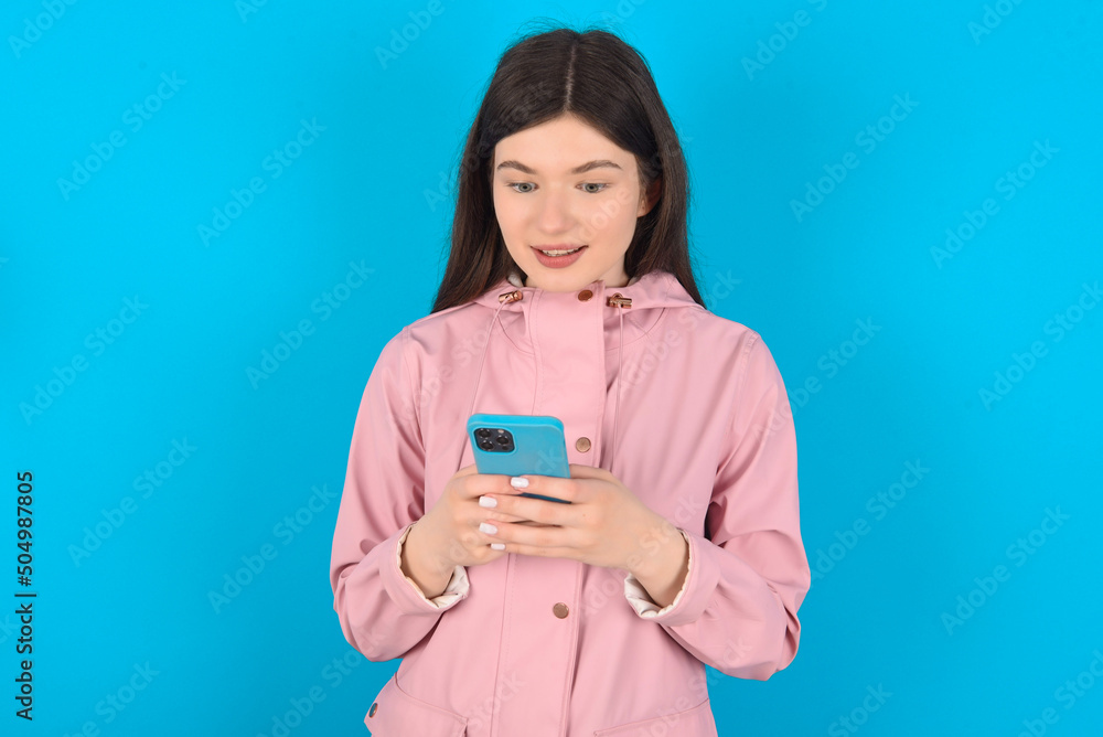young beautiful Caucasian woman wearing pink raincoat over blue wall using mobile phone chatting free time . young beautiful Caucasian woman wearing pink raincoat over blue wall using mobile phone chatting free time .