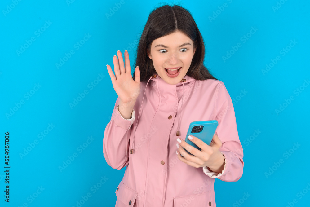 young beautiful Caucasian woman wearing pink raincoat over blue wall holding in hands cell reading browsing news young beautiful Caucasian woman wearing pink raincoat over blue wall holding in hands cell reading browsing news