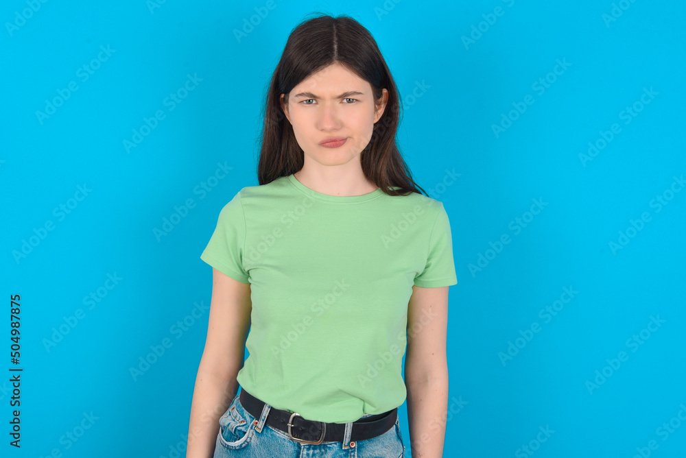 Offended dissatisfied young beautiful Caucasian woman wearing green T-shirt over blue wall with moody displeased expression at camera being disappointed by something Offended dissatisfied young beautiful Caucasian woman wearing green T-shirt over blue wall with moody displeased expression at camera being disappointed by something