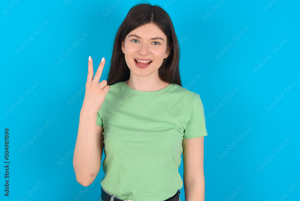 Fototapeta premium young beautiful Caucasian woman wearing green T-shirt over blue wall smiling and looking friendly, showing number two or second with hand forward, counting down
