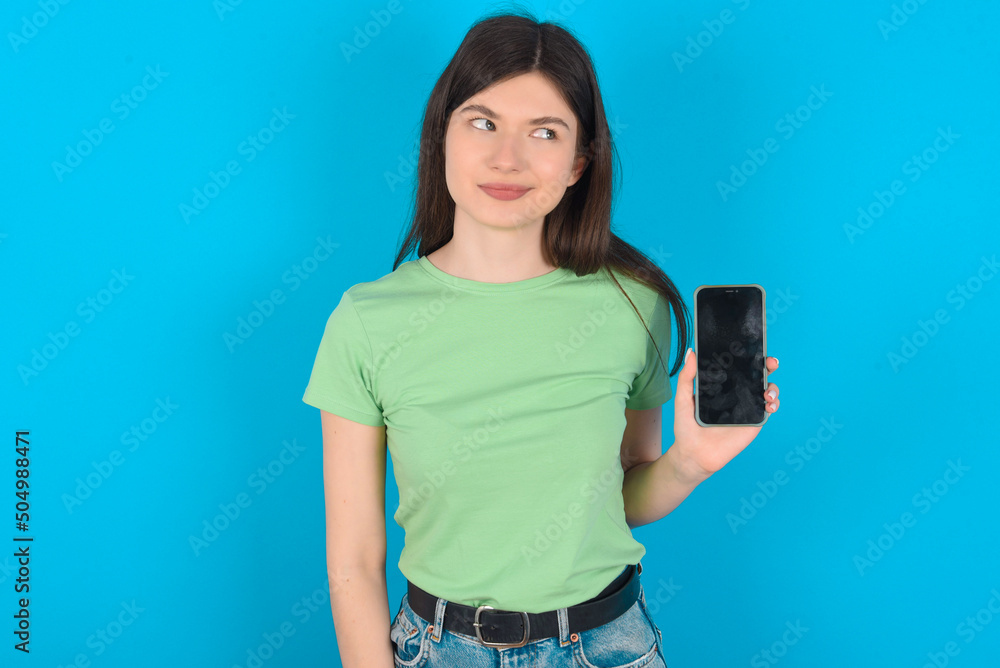young beautiful Caucasian woman wearing green T-shirt over blue wall holds new mobile phone and looks mysterious aside shows blank display of modern cellular young beautiful Caucasian woman wearing green T-shirt over blue wall holds new mobile phone and looks mysterious aside shows blank display of modern cellular