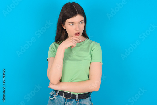 Astonished young beautiful Caucasian woman wearing green T-shirt over blue wall looks aside surprisingly with opened mouth. © Roquillo