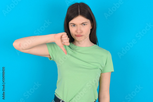 young beautiful Caucasian woman wearing green T-shirt over blue wall looking unhappy and angry showing rejection and negative with thumbs down gesture. Bad expression.