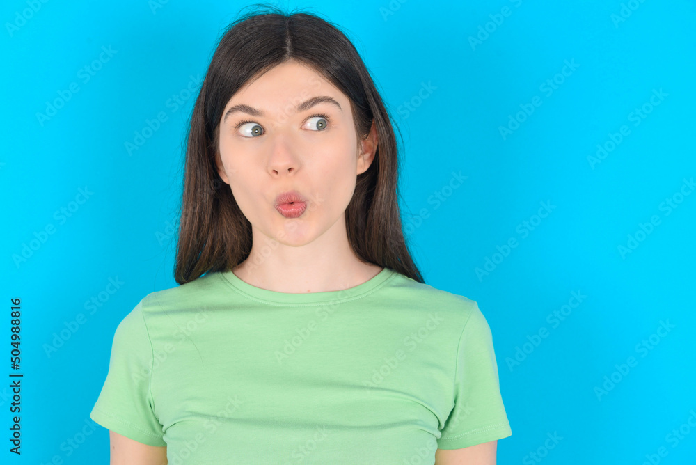 Fototapeta premium Shocked young beautiful Caucasian woman wearing green T-shirt over blue wall look empty space with open mouth screaming: Oh My God! I can't believe this.