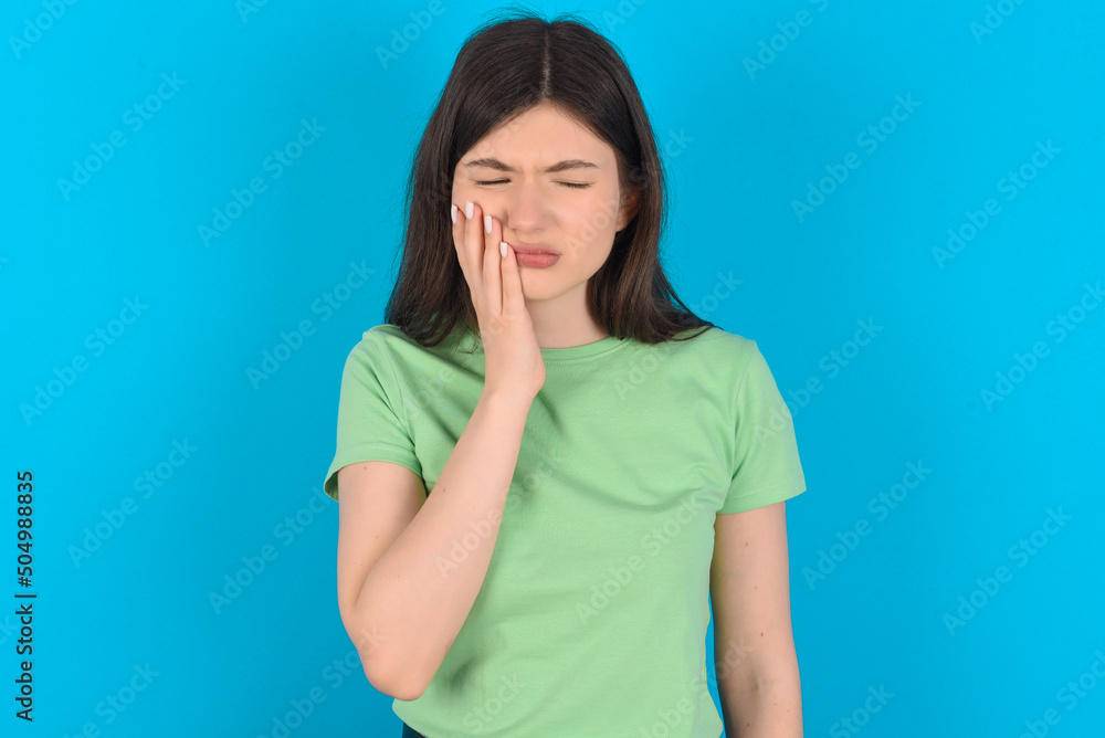 young beautiful Caucasian woman wearing green T-shirt over blue wall with toothache young beautiful Caucasian woman wearing green T-shirt over blue wall with toothache