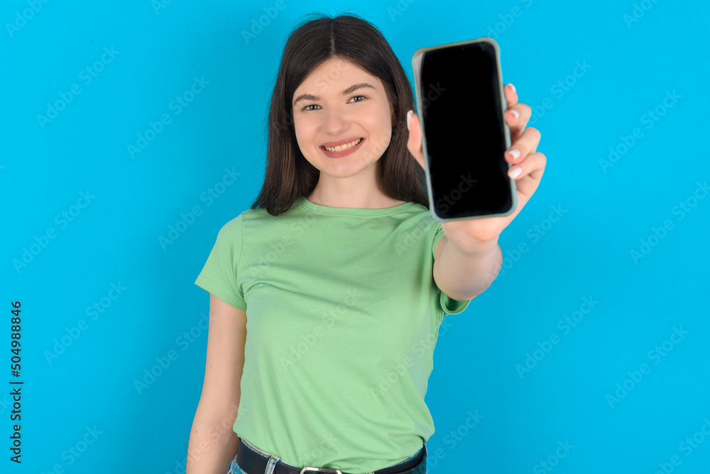 Charming adorable young beautiful Caucasian woman wearing green T-shirt over blue wall holding modern device, showing black screen smartphone