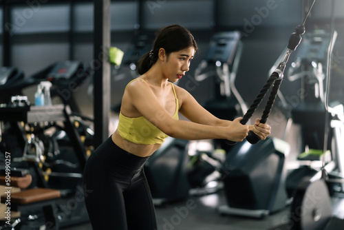 Young Asian woman flexing arm muscles on cable machine at gym.