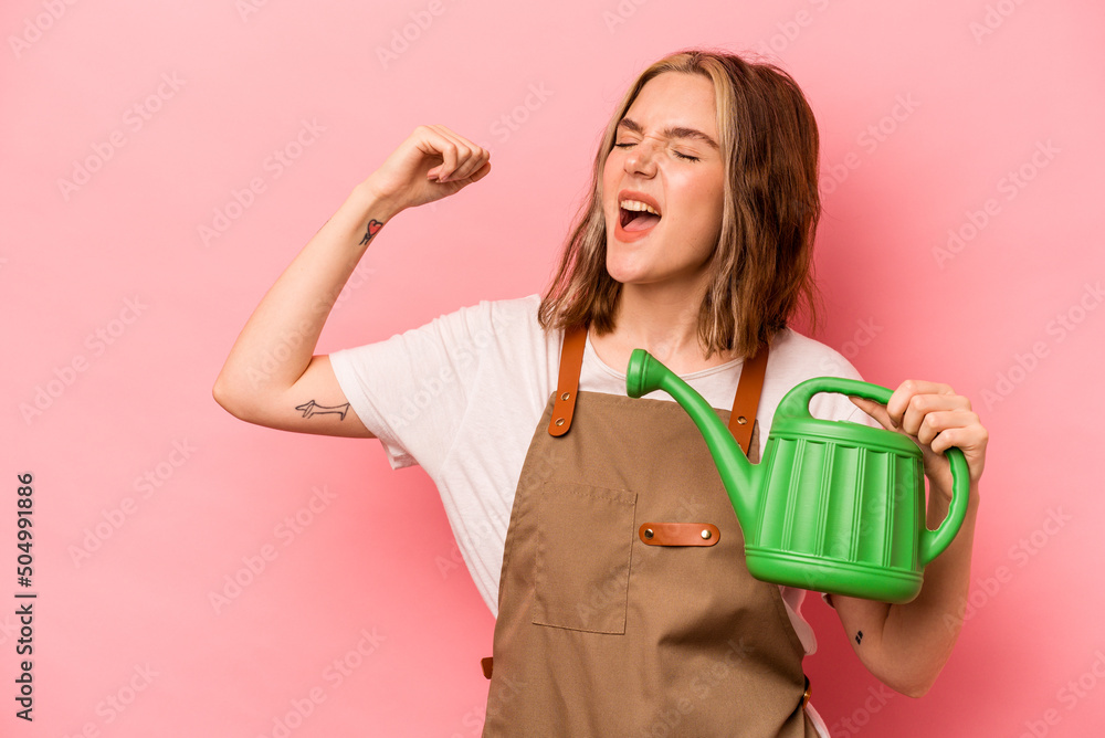Young gardener woman holding watering can isolated on pink background raising fist after a victory, winner concept. Young gardener woman holding watering can isolated on pink background raising fist after a victory, winner concept.