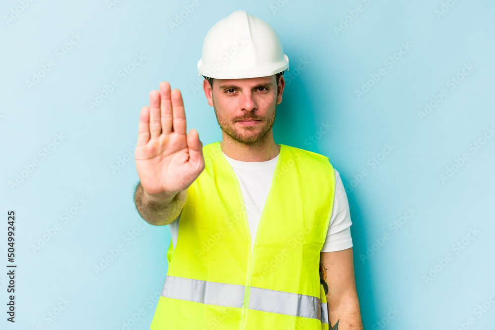Young laborer caucasian man isolated on blue background standing with outstretched hand showing stop sign, preventing you.