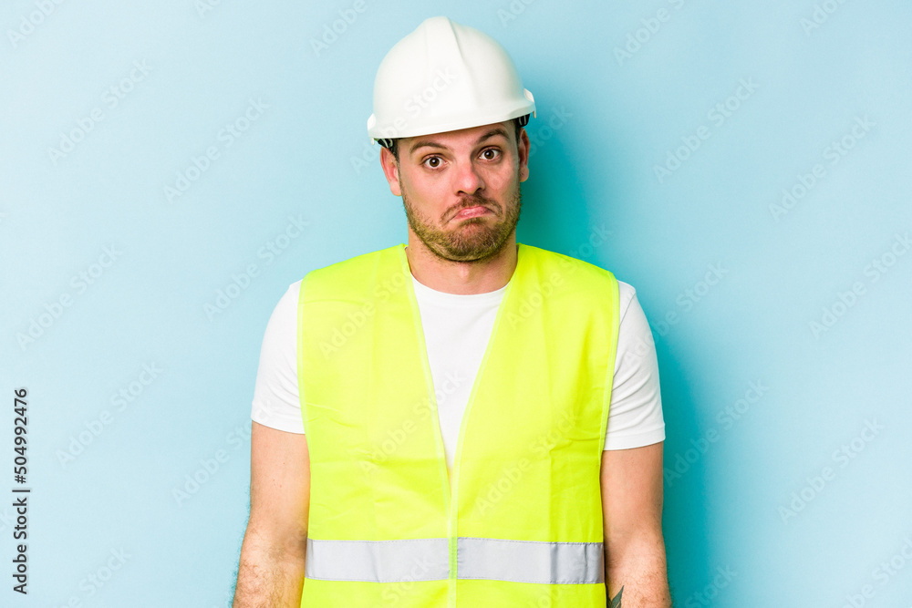 Young laborer caucasian man isolated on blue background shrugs shoulders and open eyes confused.
