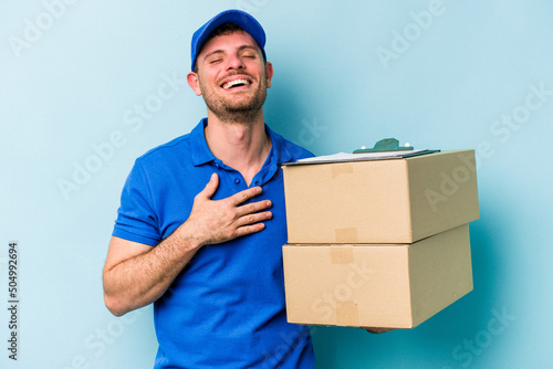 Young caucasian delivery man isolated on blue background laughs out loudly keeping hand on chest.