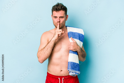 Young caucasian man holding beach towel isolated on blue background keeping a secret or asking for silence.