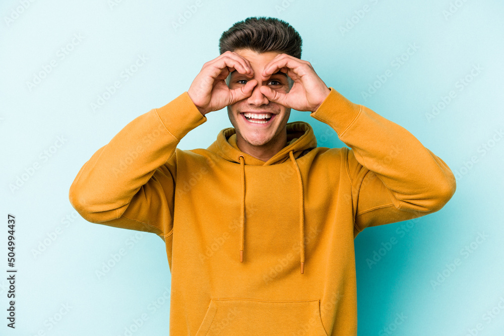 Young caucasian man isolated on blue background showing okay sign over eyes Young caucasian man isolated on blue background showing okay sign over eyes