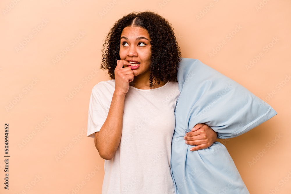 Young African American woman holding a pillow isolated on beige background relaxed thinking about something looking at a copy space. Young African American woman holding a pillow isolated on beige background relaxed thinking about something looking at a copy space.