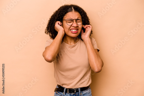Young African American woman isolated on beige background covering ears with hands.