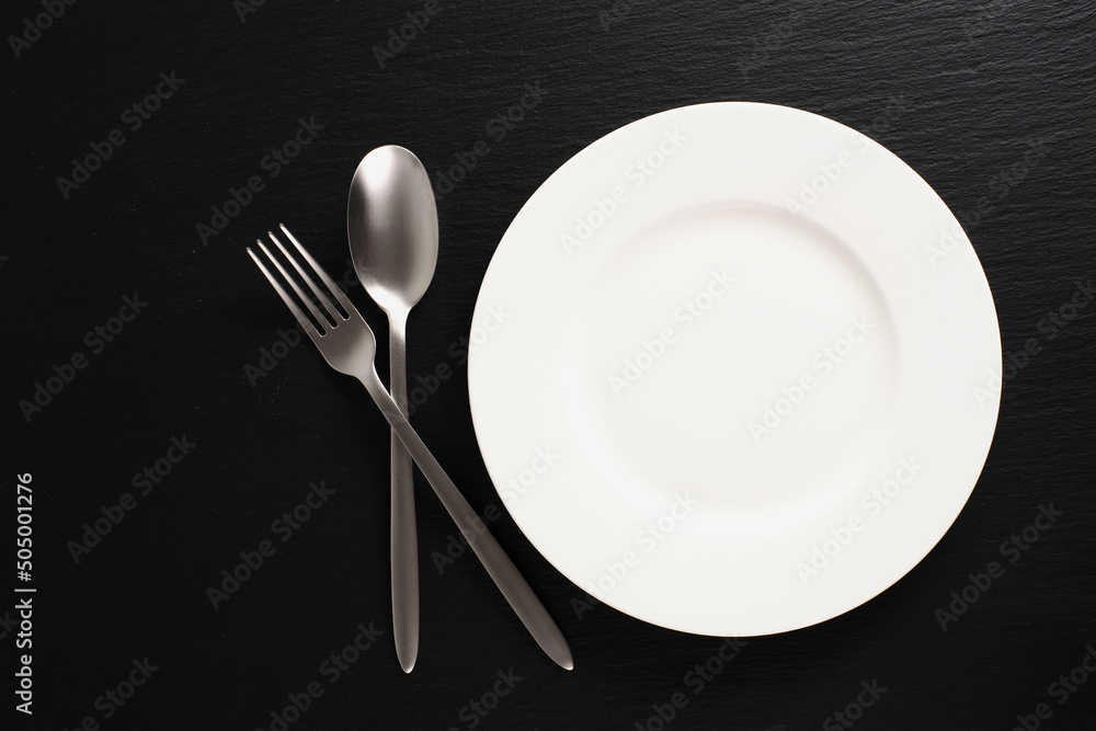 White plate on a black background. Empty white plate, fork, spoon on black slate, flat lay. White plate on a black background. Empty white plate, fork, spoon on black slate, flat lay.