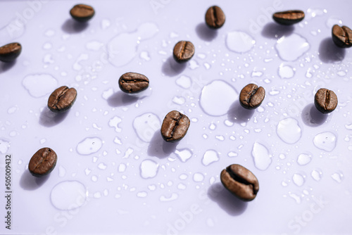 Coffee beans on a light lilac surface next to water drops. Coffee background. Coffee advertisement. Side view.