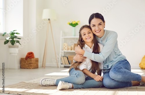 Portrait of a happy beautiful mother and her child at home. Young mom and her pretty little daughter sitting on the floor in a modern interior of their own apartment, smiling and looking at the camera photo