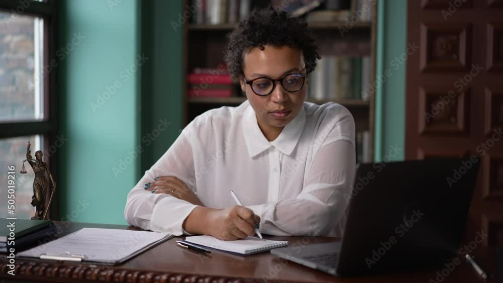 4k Young businesswoman looking at laptop and writing in notebook, sitting at table in company spbas. Portrait beautiful american african woman looks at computer screen and writes on paper, works or 4k Young businesswoman looking at laptop and writing in notebook, sitting at table in company spbas. Portrait beautiful american african woman looks at computer screen and writes on paper, works or