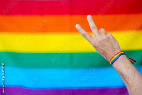 Mano haciendo el símbolo de la paz con pulsera de colores del colectivo lgtbi, con la bander lgtbi de fondo photo