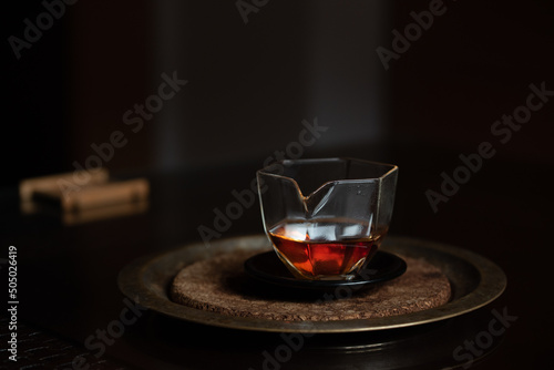 A small Chinese glass jug stands on a dark table against a dark background. There is caramel-colored tea in the pot. The pot is on a tray.