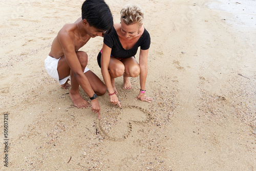 Multiracial couple drawing a heart on the sand of a tropical beach
