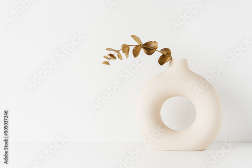 Round vase with a golden-colored eucalyptus branch on a white table by the wall.