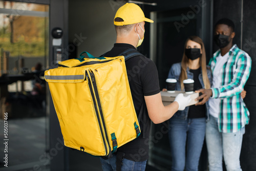 Cacasian man in face mask works on the delivery of pizza. Young multiracial friends, ordered the delivery of pizza. Pizza deliveryman brought order to house working during covid-19 photo