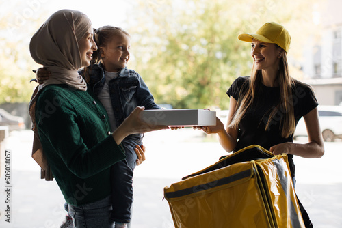 Smiling delivery woman in yellow uniform cap delivering cardboard box with pizza and coffee to attractive islamic woman with little daughter recipient. Courier service concept.