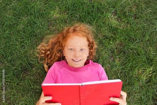 Little redhead girl reading book on green grass photo