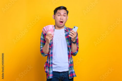 Excited young Asian man in plaid shirt holding mobile phone and money banknotes, got easy loan isolated on yellow background
