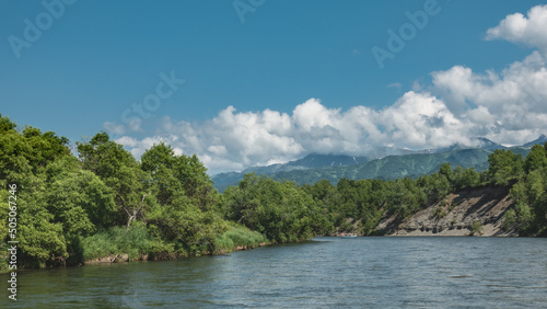 The river flows calmly between steep banks overgrown with forest. An inflatable rafting boat with people is visible in the distance. A mountain range against a blue sky and clouds. Kamchatka