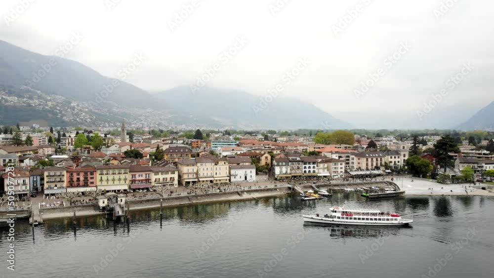 Aerial flyover alongside the waterfront of Ascona in Ticino, Switzerland with a view of a boat arriving at the pier and the city buildings along the lakeside promenade on the shores of Lago Maggiore Aerial flyover alongside the waterfront of Ascona in Ticino, Switzerland with a view of a boat arriving at the pier and the city buildings along the lakeside promenade on the shores of Lago Maggiore