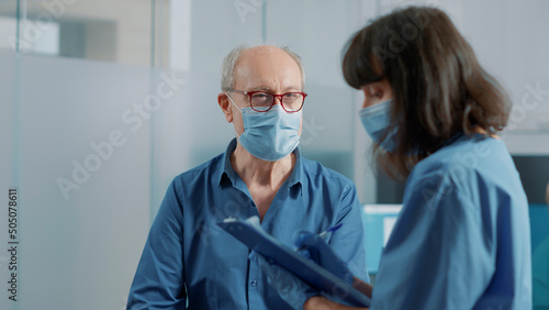 Elder patient attending checkup examination with nurse in office, talking about alternative medicine and treatment. Health assistant taking notes and discussing with old man during pandemic.